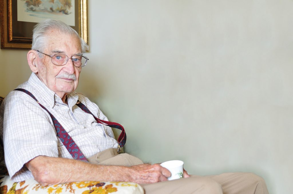 Older man with short grey hair and red suspenders sits in chair holding tea