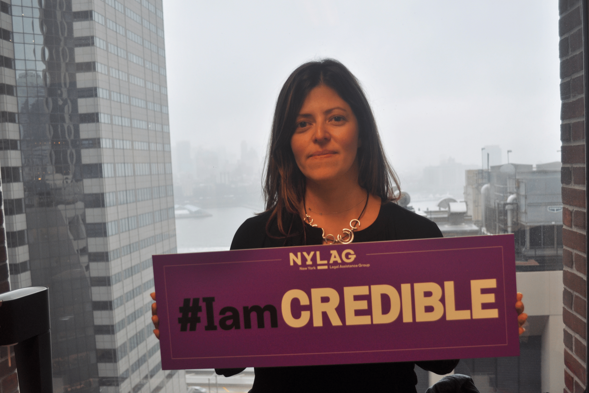 Lisa Rivera, NYLAG's Managing Editor, stand in front of a view of the East River and Brooklyn. She is holding a purple sign that says hashtag I am Credible