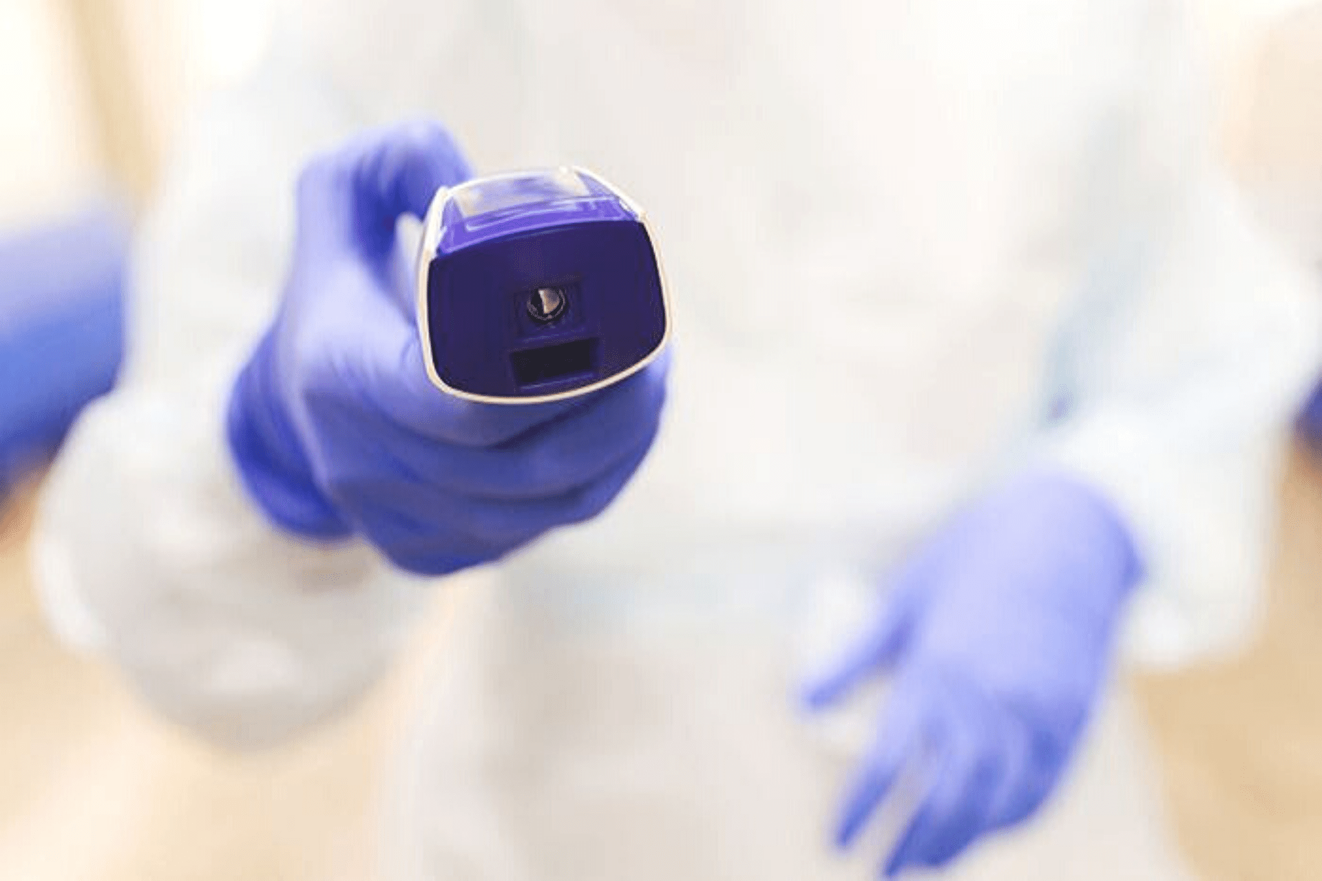 Image of a contact-less thermometer held by a medical professional with purple gloves on.