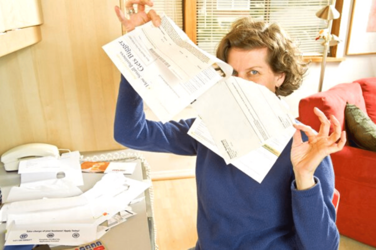 A woman sitting in her home office. She looks relieved as she rips up a bill from a big stack on her desk.