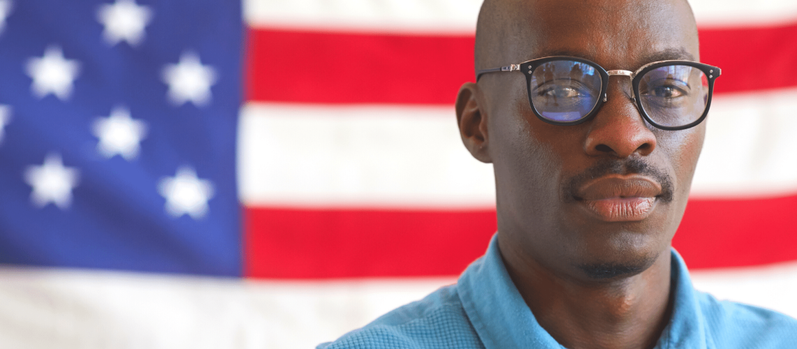 Black man wearing glasses with the American flag behind him.